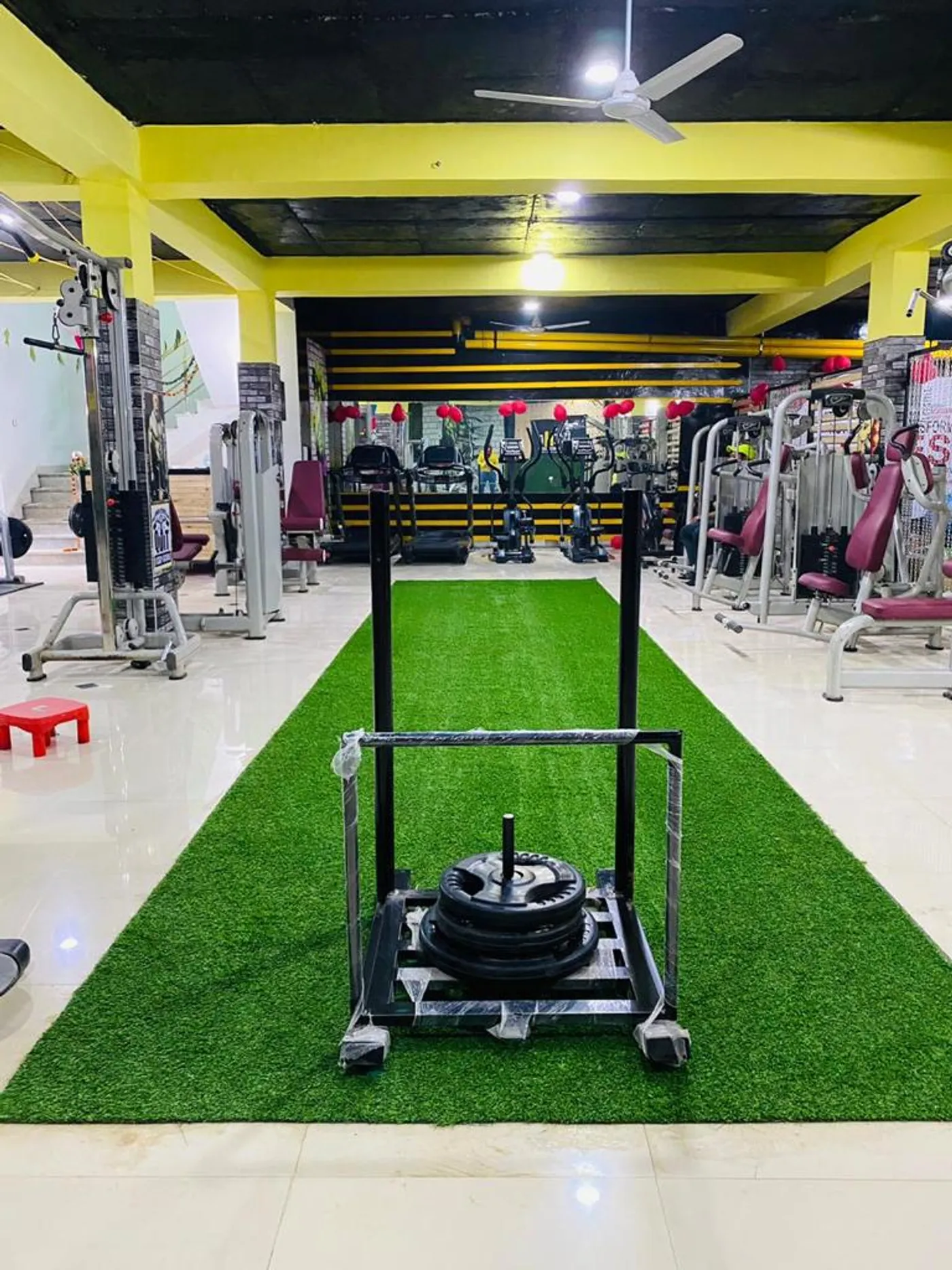 A long artificial-turf lane down the centre of the gym floor, a weighted sled at one end, with exercise machines lined up on both sides and a staircase visible at the far end.