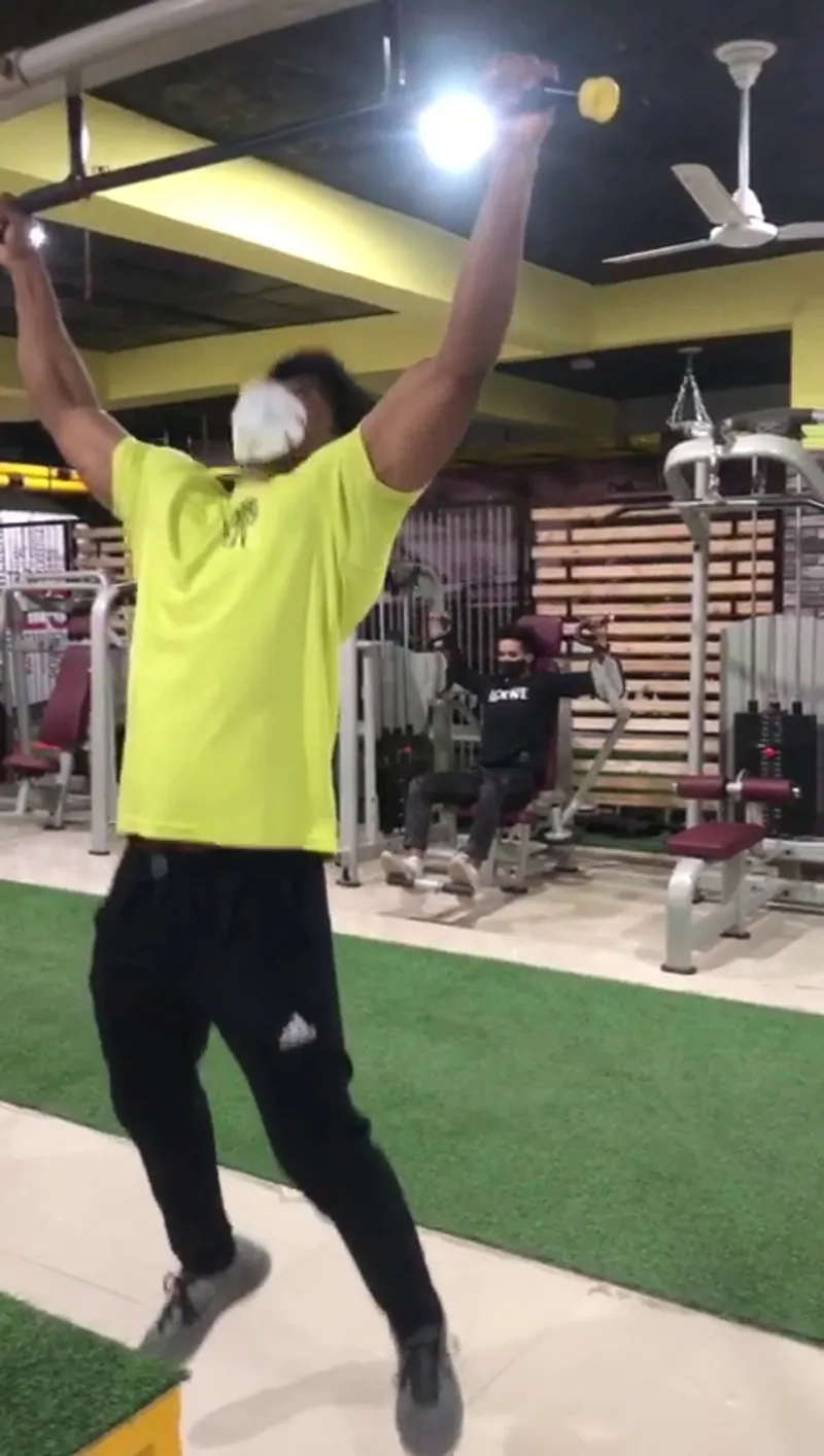 A member mid-exercise on a pull-up bar, another person exercising on a seated machine behind him, on the artificial-turf training lane.